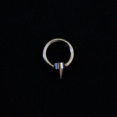 piercing captive cone da tal da realeza em um fundo preto aveludado. a peça é prateada, tem formato circular e possui um detalhe em formato de cone.