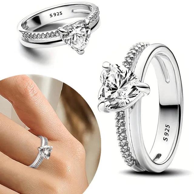 Anel de prata 925 com pedra transparente em formato de coração em um fundo branco. Também é possível observar o anel sendo usado no dedo anelar de uma mulher de pele clara.