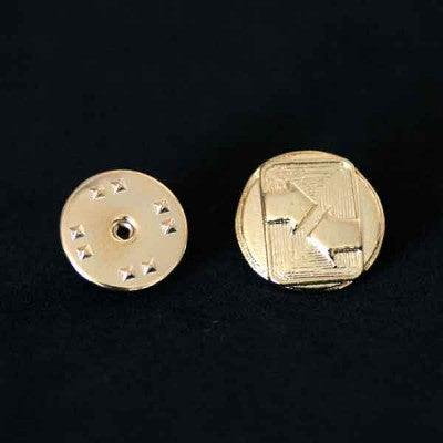 Broche Comunicação folheado a ouro em um fundo preto. A peça tem formato de setas horizontais que apontam uma para a outra.
