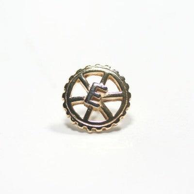Broche Engenharia Mecânica folheado a ouro em um fundo branco. A peça tem formato de engrenagem, com a letra "E" no centro.