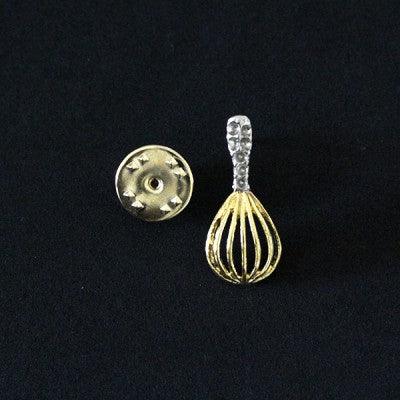 Broche Comunicação folheado a ouro em um fundo preto. A peça tem formato de um fouet, instrumento usado para bater claras.