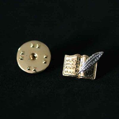 Broche Letras folheado a ouro em um fundo preto. A peça tem o formato de um livro aberto com uma pena no centro.