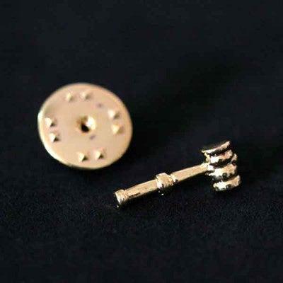Broche Direito folheado a ouro em um fundo preto. A peça tem formato de um malhete, o martelo de madeira utilizado pelos juízes.