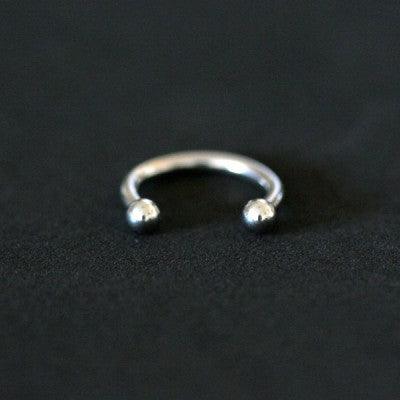 Piercing Barbell da Tal da Realeza em um fundo preto aveludado. A peça é prateada, tem formato de ferradura e possui esferas nas extremidades.