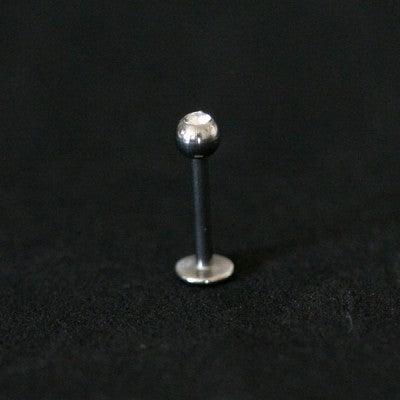 Piercing labret da Tal da Realeza em um fundo preto aveludado. A peça é prateada e possui uma esfera com pedra de zircônia na extremidade.