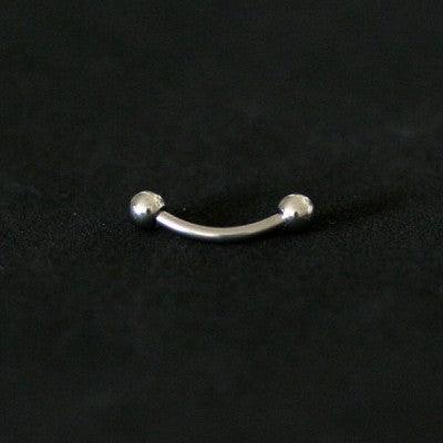 Piercing de aço cirúrgico com haste curva e esferas nas extremidades sobre fundo preto.