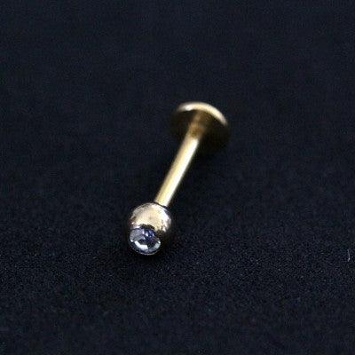 Piercing labret da Tal da Realeza em um fundo preto aveludado. A peça é dourada e possui uma esfera com pedra de zircônia na extremidade.