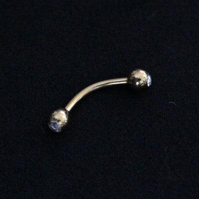 Piercing de aço cirúrgico banhado a ouro com haste curva e esferas nas extremidades sobre fundo preto. Há pedras de zircônia nas esferas.