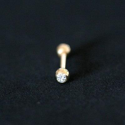 Piercing de aço cirúrgico banhado a ouro com haste reta e esferas nas extremidades sobre fundo preto. Há uma pedra de cristal em uma das esferas.
